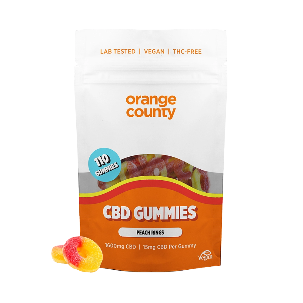 An orange and white stand-up pouch of Orange County CBD Gummies, labeled 'Peach Rings', with two gummy candies placed in front on a white background.