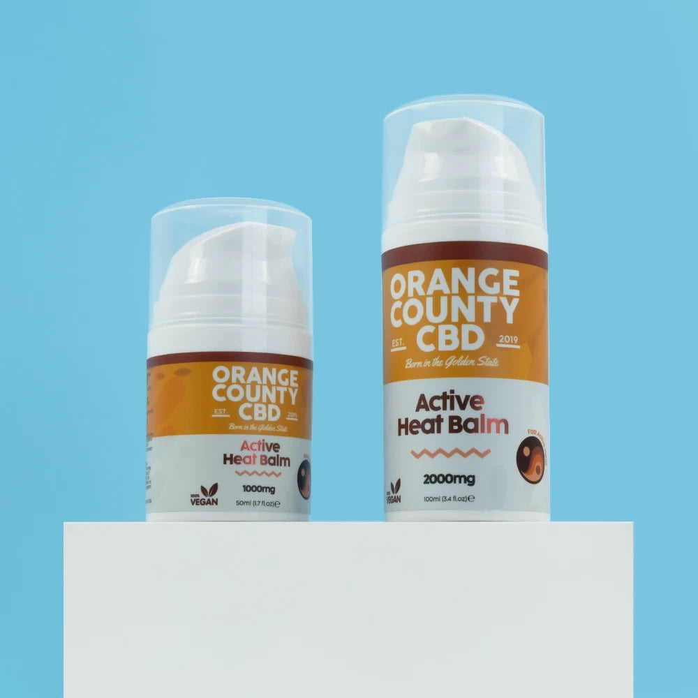 Two clear plastic pump bottles of Orange County CBD Active Heat Balm, one 1000mg and one 2000mg, displayed on a white pedestal against a solid light blue background.