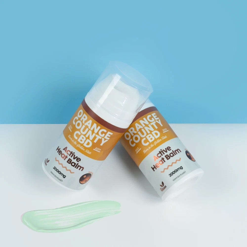 Two bottles of Orange County CBD Active Heat Balm 300mg are shown on a white surface against a light blue background, with a light green balm smear in the foreground.