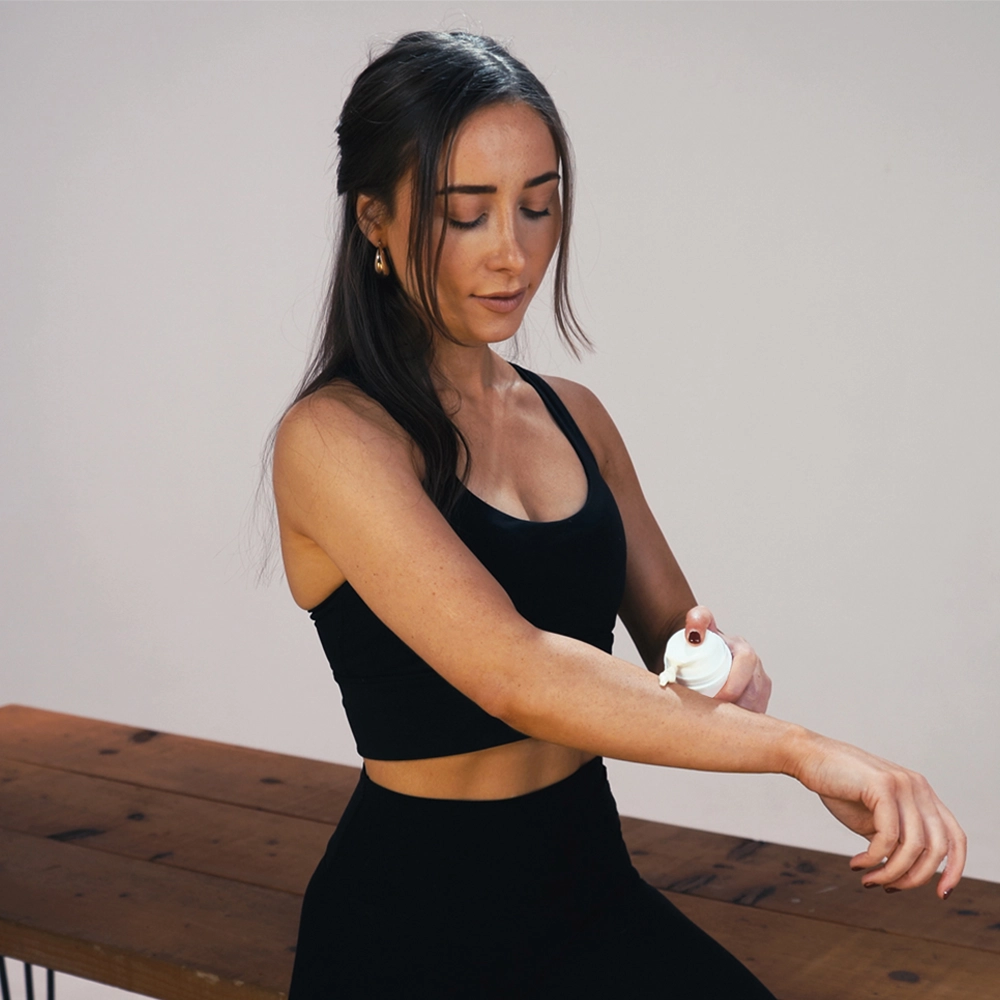 A woman in a black sports bra and leggings applies a white cream from a small pump bottle onto her forearm, seated on a wooden bench against a plain light gray wall.