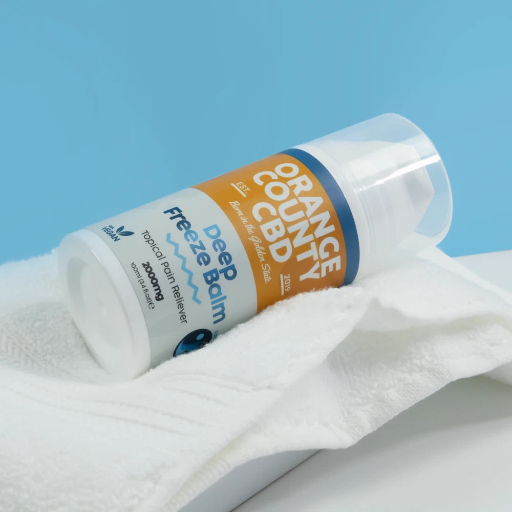 A bottle of Freeze Balm by Orange County CBD lies on a white towel against a light blue background. The product label is white and orange with blue text and accents.