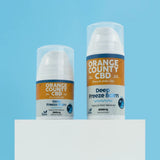 Two topical pain reliever balm containers from Orange County CBD on a white pedestal against a light blue background. The smaller container is Deep Freeze Balm 1000mg, and the larger container is Deep Freeze Balm 2000mg, both with white caps and clear plastic tops.