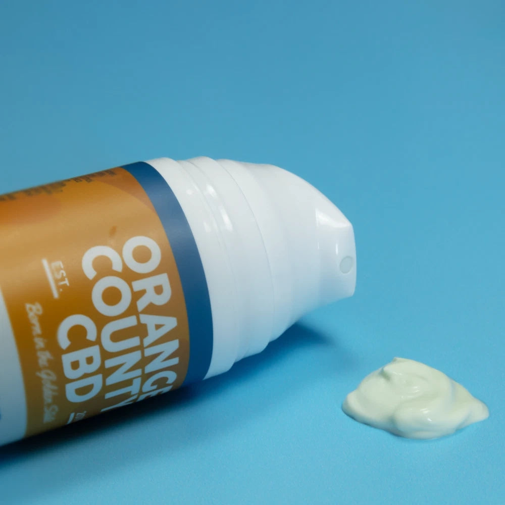 A squeeze tube of CBD cream lies on its side on a bright blue background, with a dollop of pale yellow cream spilled in front. The tube is white and orange with dark blue accents.