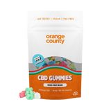 An orange and white stand-up pouch of Orange County CBD Gummies, shown on a clean white background with three loose gummies in front. The packaging features a clear window displaying multicolored gummies and includes text such as 'LAB TESTED | VEGAN | THC-FREE', '100 GUMMIES', and 'CBD GUMMIES'.