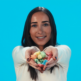 A smiling woman with long dark hair holds a small pile of colorful, CBD gummy bears toward the camera against a bright cyan background.