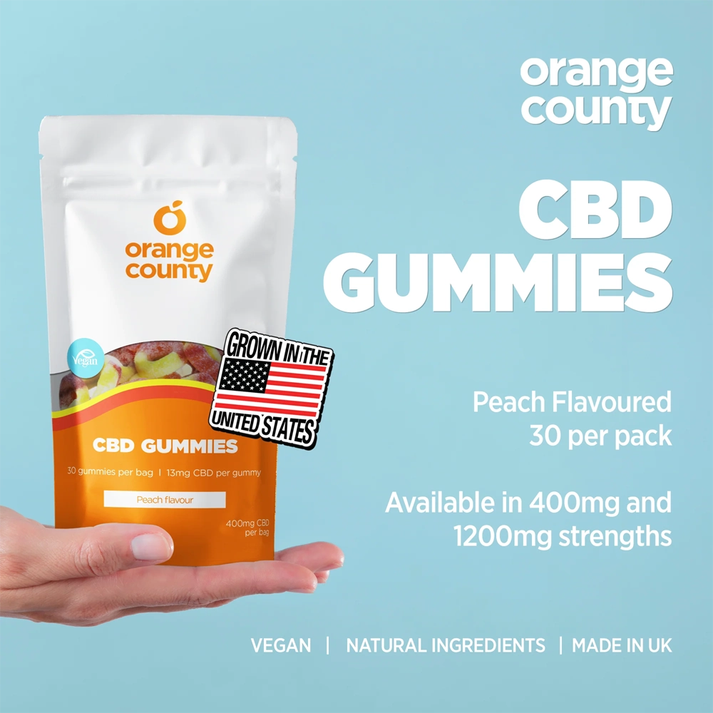 A hand holding an Orange County CBD Gummies pouch against a light blue background with promotional text. The pouch is peach and white with a fruit image on the label and a 'Grown in the United States' badge.