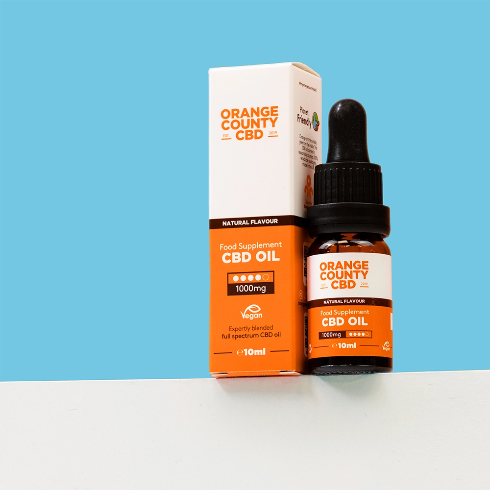 An amber glass dropper bottle of Orange County CBD Oil next to its white and orange cardboard packaging box, displayed on a white surface against a solid blue background.
