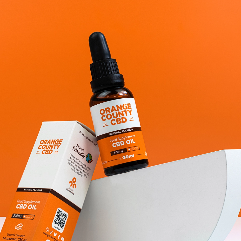 An amber glass dropper bottle of Orange County CBD food supplement CBD oil stands next to its white and orange retail box on a white curved riser, set against a solid orange background.