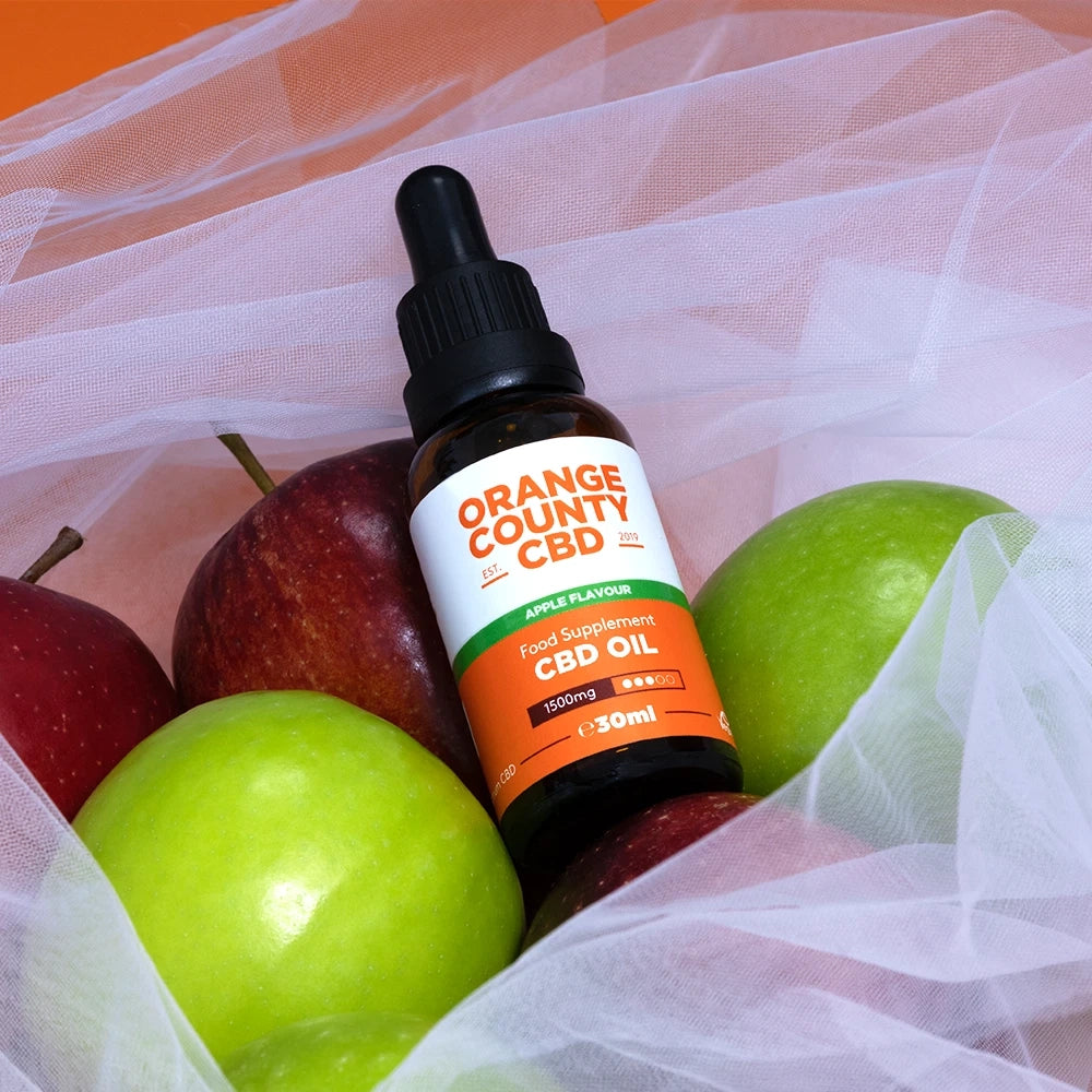 A 30ml bottle of Orange County CBD Apple Flavour food supplement CBD oil, styled among fresh red and green apples inside a white tulle bag.
