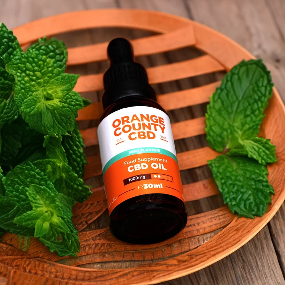 A 30ml bottle of Orange County CBD Oil in the Mint Leaf flavor, displayed on a round wooden tray with fresh mint leaves on a rustic wooden table.