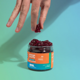 A hand is reaching for dark red gummy candies from a clear jar labeled 'orange county' and 'NMN' against a bright turquoise background.