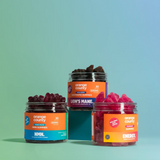 Three jars of Orange County gummy candies arranged on a teal-to-light-blue gradient background. The jars are labeled NMM, Lion's Mane, and Energy, showing dark red, pink, and brown gummies inside clear plastic containers with orange and blue labels.